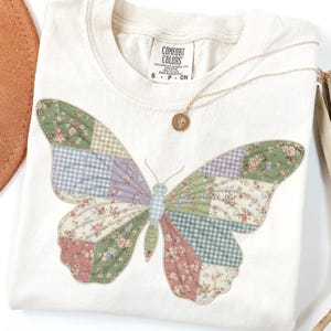 以下が含まれることがあります： クリーム色のTシャツ。緑、ピンク、紫の色合いのパッチワークの蝶のデザインが特徴です。蝶は花柄です。小さなペンダントが付いたゴールドのネックレスがシャツの上にあります。「COMFORT COLORS」と書かれたタグが付いています。