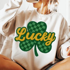 K&ouml;nnte beinhalten: Cremefarbenes Sweatshirt mit einem gr&uuml;nen und gelben Kleeblatt-Design. Das Kleeblatt ist ein dreibl&auml;ttriges Kleeblatt mit einem karierten Muster, und das Wort "Lucky" ist in gelber, kursiver Schrift &uuml;ber dem Kleeblatt geschrieben. Das Sweatshirt ist ein l&auml;ssiger Stil.