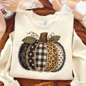 May include: Cream-colored sweatshirt featuring a patchwork pumpkin design. The pumpkin has sections of black lace, polka dots, buffalo plaid, and leopard print. The pumpkin is topped with a brown stem and green leaves, perfect for fall.