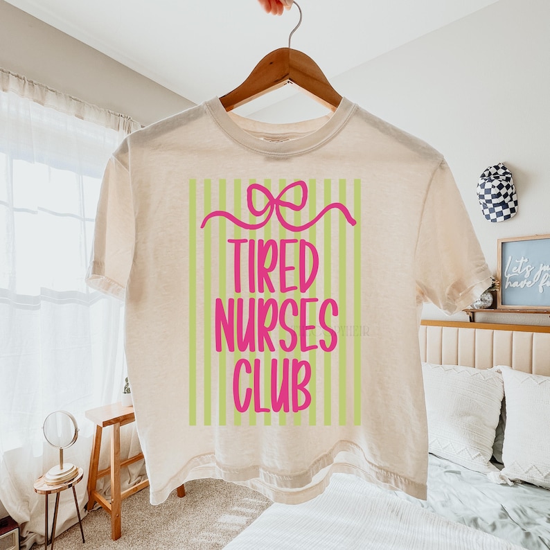 May include: A cream-colored t-shirt with the text "TIRED NURSES CLUB" in pink, set against a green and white striped background. A pink bow design is above the text. The shirt is hanging on a wooden hanger.