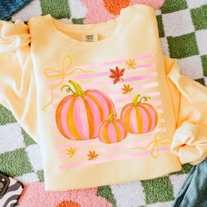 May include: A light yellow sweatshirt featuring a fall-themed design. The design includes three pink and orange pumpkins, pink horizontal stripes, yellow bows, and autumn leaves. The sweatshirt is folded and displayed on a checkered surface.