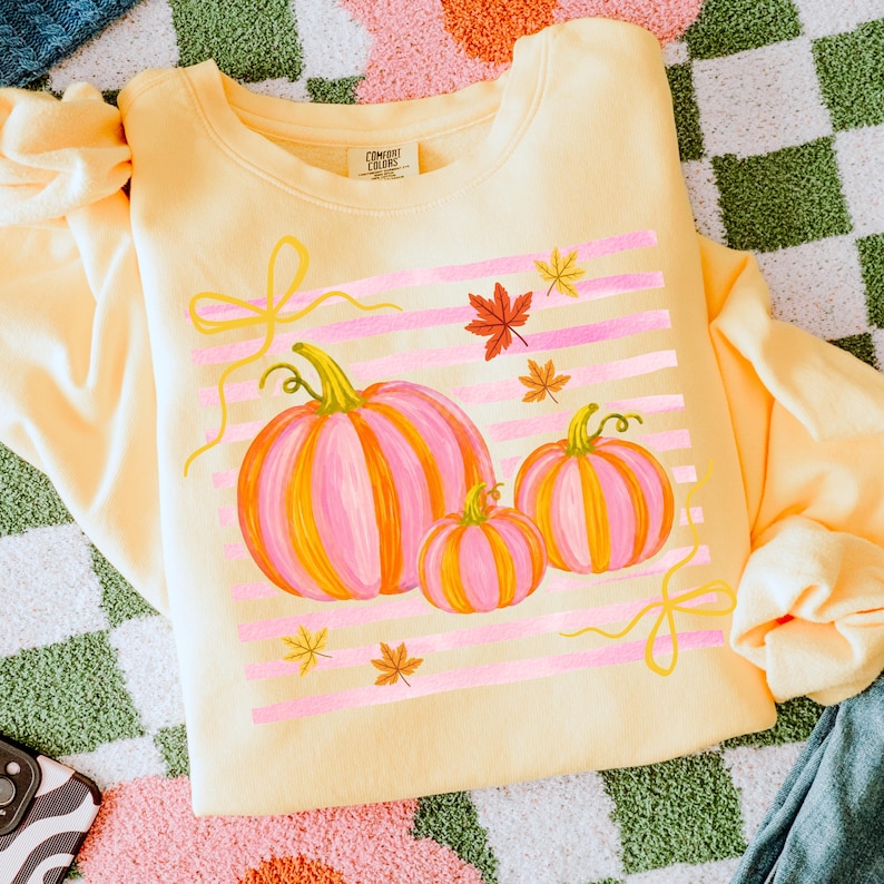 May include: A light yellow sweatshirt featuring a fall-themed design. The design includes three pink and orange pumpkins, pink horizontal stripes, yellow bows, and autumn leaves. The sweatshirt is folded and displayed on a checkered surface.