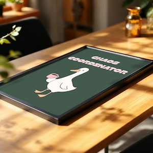 May include: A framed art print with a white cartoon goose holding a pink mug, and the words "CHAOS COORDINATOR" in pink text on a dark green background. The artwork is in a black frame and rests on a wooden table.