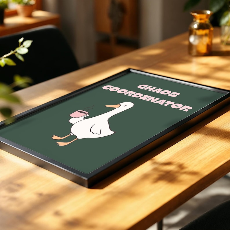 May include: A framed art print with a white cartoon goose holding a pink mug, and the words "CHAOS COORDINATOR" in pink text on a dark green background. The artwork is in a black frame and rests on a wooden table.