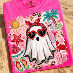 May include: A hot pink Comfort Colors t-shirt featuring a whimsical ghost wearing heart-shaped sunglasses and a red bandana. The design includes a cocktail, palm tree, starfish, watermelon, and other summer-themed elements.