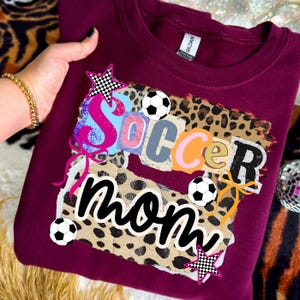 May include: Burgundy sweatshirt with a colorful "Soccer Mom" graphic. The design includes soccer balls, stars, and a leopard print background. The text is in a collage style with various fonts and colors.
