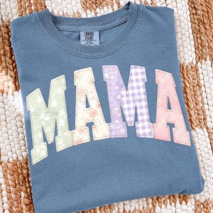 May include: A blue Comfort Colors sweatshirt with the word "MAMA" in a patchwork design. The letters feature floral, gingham, and solid patterns in pastel colours. The sweatshirt has a crew neck and a tag that says "Comfort Colors".