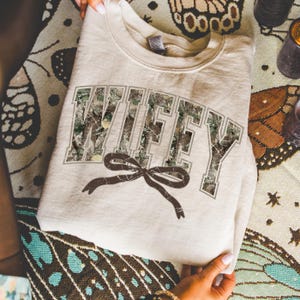 May include: A beige sweatshirt with the word "WIFEY" in a camouflage pattern across the front. A brown bow is printed below the word. The sweatshirt is folded and resting on a patterned surface.