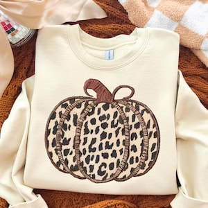 Peut inclure: Sweat-shirt crème avec un grand motif de citrouille. La citrouille a une tige marron et un motif léopard noir et marron. Le sweat-shirt est présenté sur une couverture en tricot marron.