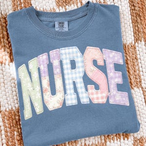 May include: A blue Comfort Colors t-shirt with the word "NURSE" appliquéd across the front. The letters feature floral and gingham patterns, with a stitched outline. The shirt is displayed on a woven surface.