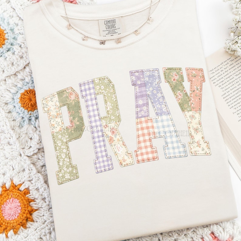 K&ouml;nnte beinhalten: Cremefarbenes Sweatshirt mit dem Wort "PRAY" in Patchwork-Buchstaben. Jeder Buchstabe hat ein anderes Blumen- oder Vichy-Muster in Gr&uuml;n-, Lila-, Orange- und Rosat&ouml;nen. Eine zarte Schmetterlingskette liegt auf dem Shirt.