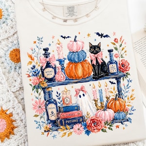 May include: A cream-colored t-shirt featuring a graphic print of two shelves filled with Halloween-themed items including a black cat with a pink bow, stacked pumpkins, potion bottles labeled Witches Brew, Magic Potion, and Poison, books, and candles.