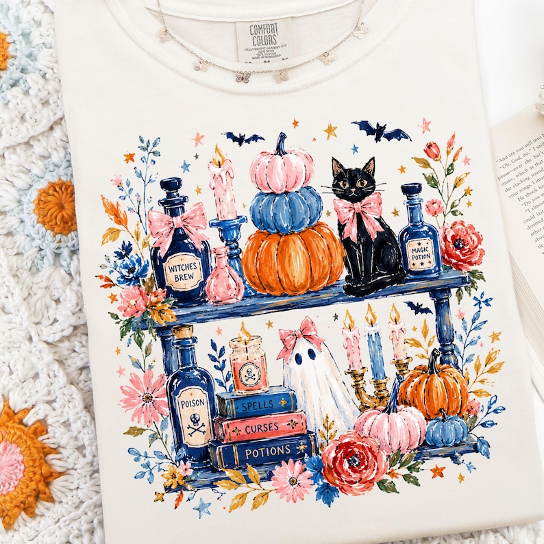 May include: A cream-colored t-shirt featuring a graphic print of two shelves filled with Halloween-themed items including a black cat with a pink bow, stacked pumpkins, potion bottles labeled Witches Brew, Magic Potion, and Poison, books, and candles.