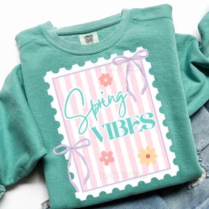 May include: A teal Comfort Colors sweatshirt with a graphic that says "Spring VIBES" in a stamp-like design. The design includes pink and white stripes, flowers, and purple bows. The sweatshirt is folded on top of denim.