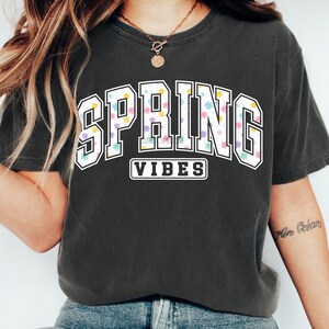 May include: Dark gray t-shirt with the words "SPRING VIBES" in a white, floral-filled design. The word "SPRING" is in a collegiate-style font, and "VIBES" is in a smaller, rectangular box below.