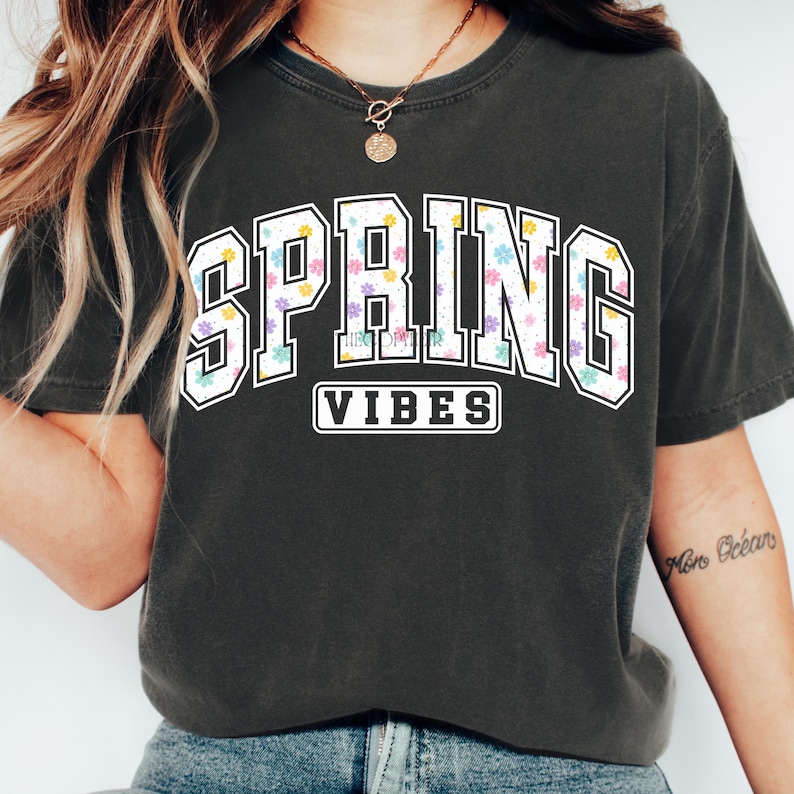 May include: Dark gray t-shirt with the words "SPRING VIBES" in a white, floral-filled design. The word "SPRING" is in a collegiate-style font, and "VIBES" is in a smaller, rectangular box below.