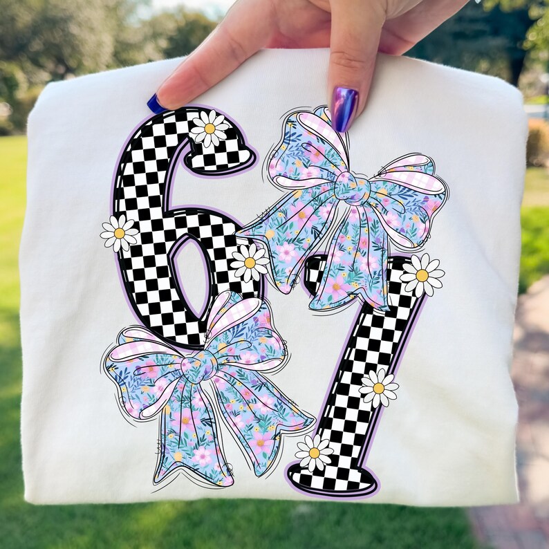 May include: Cream-colored t-shirt featuring the numbers "67" with a checkerboard pattern and floral bows. The numbers are outlined in black and purple, with white daisies scattered around them. The bows have a floral pattern with pink, blue, and yellow flowers.