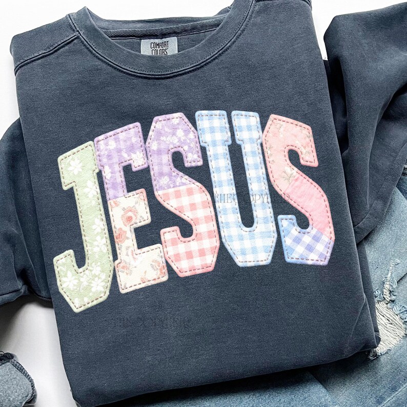 Puede incluir: Sudadera azul oscuro con la palabra "JESUS" aplicada en la parte delantera. Las letras est&aacute;n en varios patrones pastel, incluyendo florales, cuadros y colores lisos. La sudadera tiene cuello redondo y la marca "Comfort Colors" es visible.