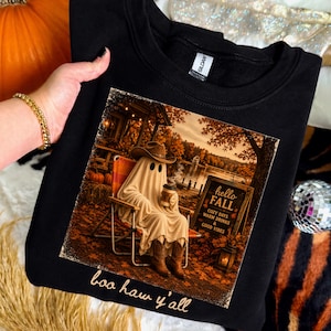 Könnte beinhalten: Schwarzes Sweatshirt mit einem Aufdruck, der einen Geist mit Cowboyhut und Stiefeln zeigt, der in einem Stuhl mit einer Kaffeetasse sitzt. Der Aufdruck enthält den Text "hello FALL" und "boo haw y'all".