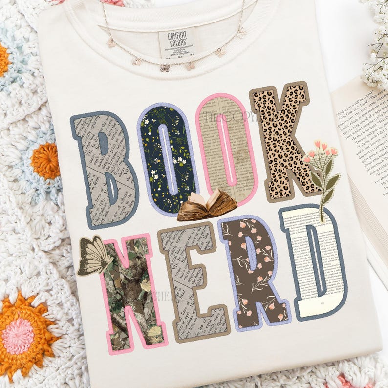 May include: Cream-colored sweatshirt with the words "BOOK NERD" in a colorful, patchwork design. The letters feature various patterns, including floral, leopard print, and book pages. A small open book and a butterfly are also included in the design.