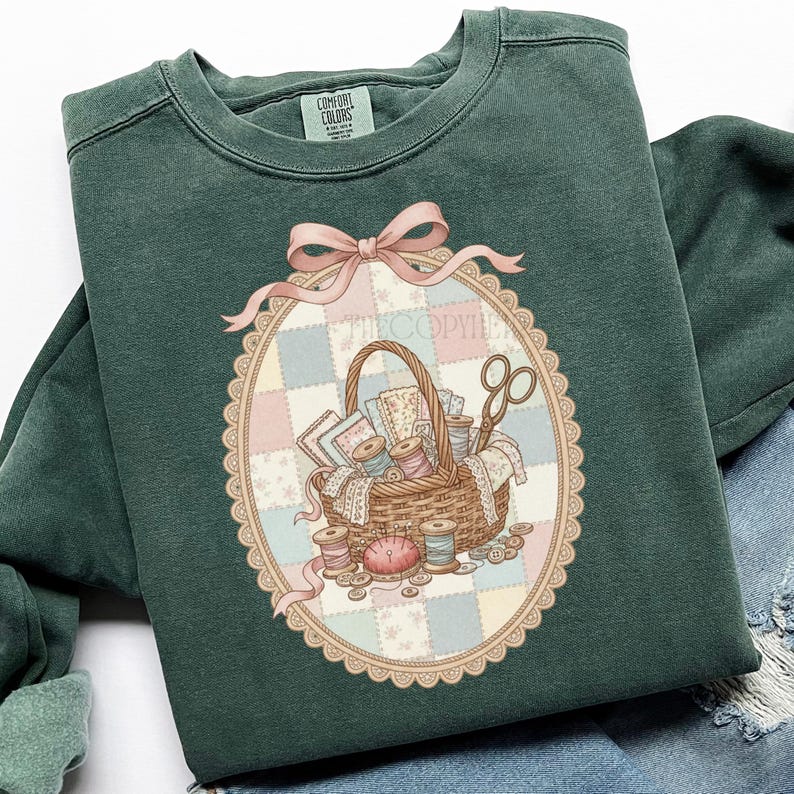 May include: A dark green sweatshirt with a detailed illustration of a sewing basket. The basket is filled with spools of thread, fabric scraps, and scissors, set against a pastel patchwork background. A pink bow and the text "THECOPYIE" are above the basket.