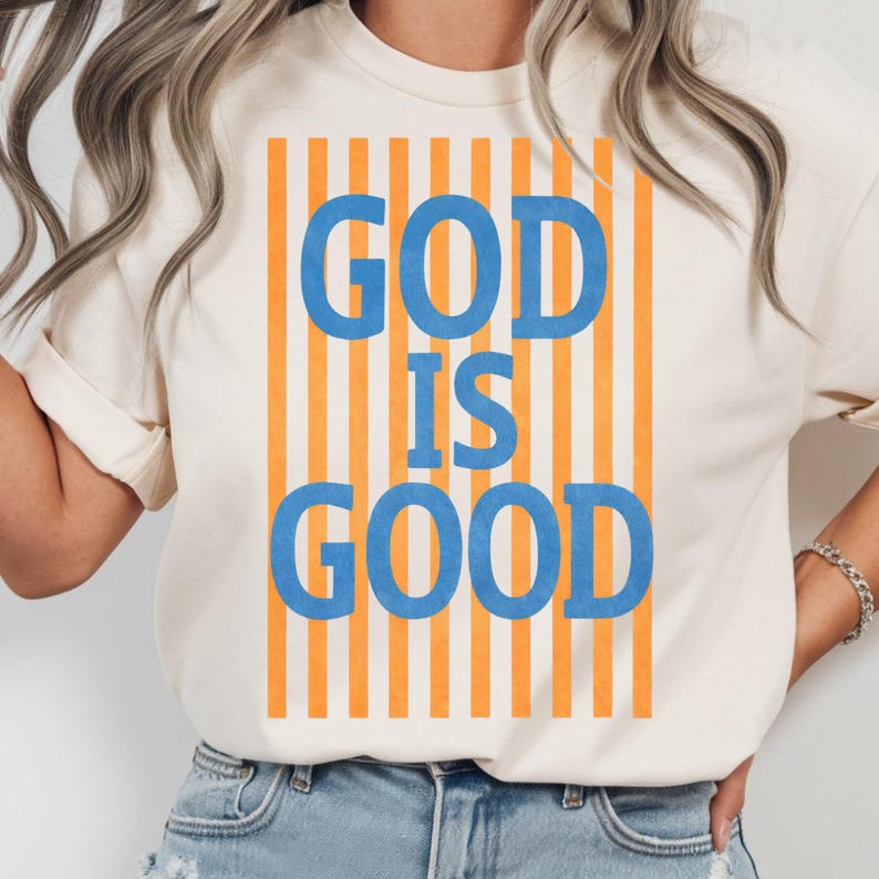 May include: Cream-colored t-shirt with the text "GOD IS GOOD" in blue letters, set against a background of vertical orange stripes. The shirt is a casual style.
