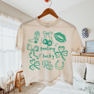 May include: Cream-colored t-shirt with green graphics, including a cowboy boot, cherries, lips, bows, clovers, dice, and a horseshoe. The words "feeling lucky" are also printed on the shirt. The shirt is hanging on a wooden hanger.