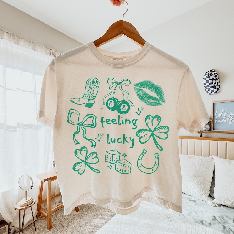 May include: Cream-colored t-shirt with green graphics, including a cowboy boot, cherries, lips, bows, clovers, dice, and a horseshoe. The words "feeling lucky" are also printed on the shirt. The shirt is hanging on a wooden hanger.