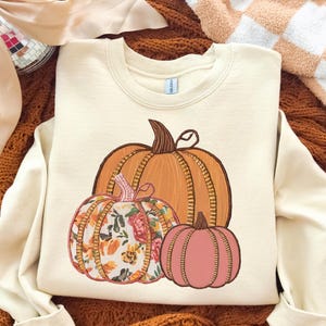Peut inclure: Sweat-shirt crème avec un motif brodé de trois citrouilles. La plus grande citrouille est orange, celle du milieu a un motif floral et la plus petite est rose. C'est un vêtement sur le thème de l'automne.