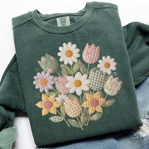 May include: A dark green sweatshirt with a floral design. The design features daisies, tulips, and other flowers in various patterns and colors. The sweatshirt has a crew neck and long sleeves. The brand tag "Comfort Colors" is visible.