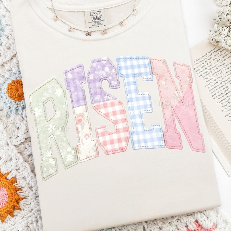 May include: Cream-colored t-shirt with the word "RISEN" in pastel patchwork letters. The letters are in green, purple, red, blue, and pink. A delicate butterfly necklace and a book are also visible.