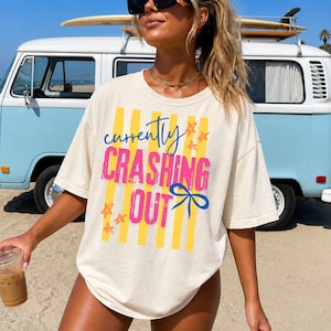 May include: Cream-colored t-shirt with the text "currently crashing out" in pink, blue, and yellow. The shirt features vertical yellow stripes and star accents. A blue and white van is in the background.