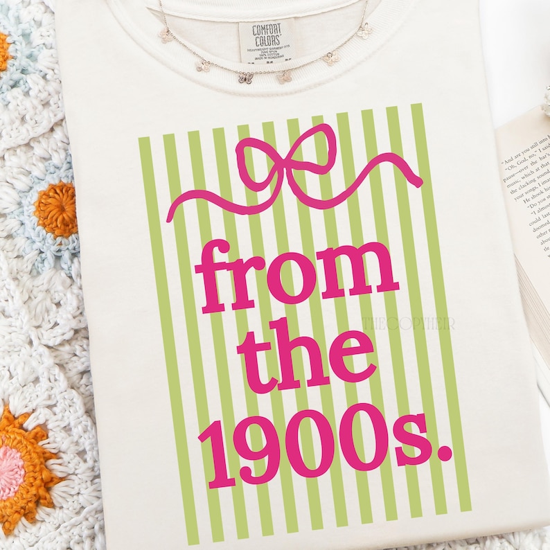 May include: Cream-colored t-shirt with a graphic design featuring a pink bow and the words "from the 1900s" in pink. The design is set against a background of vertical green stripes. A delicate butterfly necklace is visible at the top.