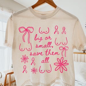 May include: Cream-colored t-shirt with pink graphics and text. The design includes illustrations of bows, flowers, and breasts, along with the words "big or small, save them all." The shirt is hanging on a wooden hanger.