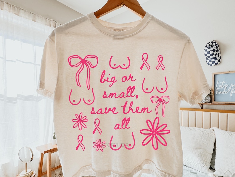 May include: Cream-colored t-shirt with pink graphics and text. The design includes illustrations of bows, flowers, and breasts, along with the words "big or small, save them all." The shirt is hanging on a wooden hanger.