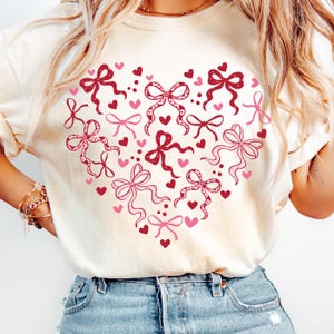 May include: Cream-colored sweatshirt featuring a heart-shaped design composed of pink and red bows and hearts. The bows vary in style and size, with some having dotted patterns. The overall design is a romantic and festive theme.