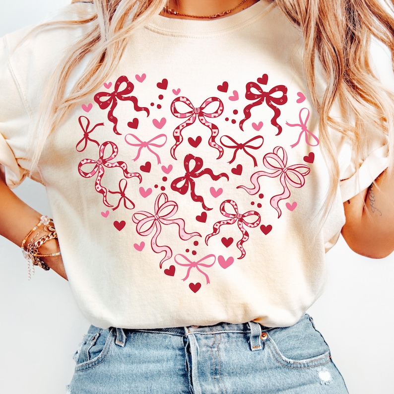 May include: Cream-colored sweatshirt featuring a heart-shaped design composed of pink and red bows and hearts. The bows vary in style and size, with some having dotted patterns. The overall design is a romantic and festive theme.
