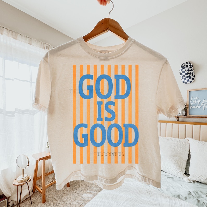 May include: Cream-colored t-shirt with the words "GOD IS GOOD" in blue block letters, set against a vertical orange striped background. The text "THECOPYHEIR" is below the main text. The shirt is hanging on a wooden hanger.