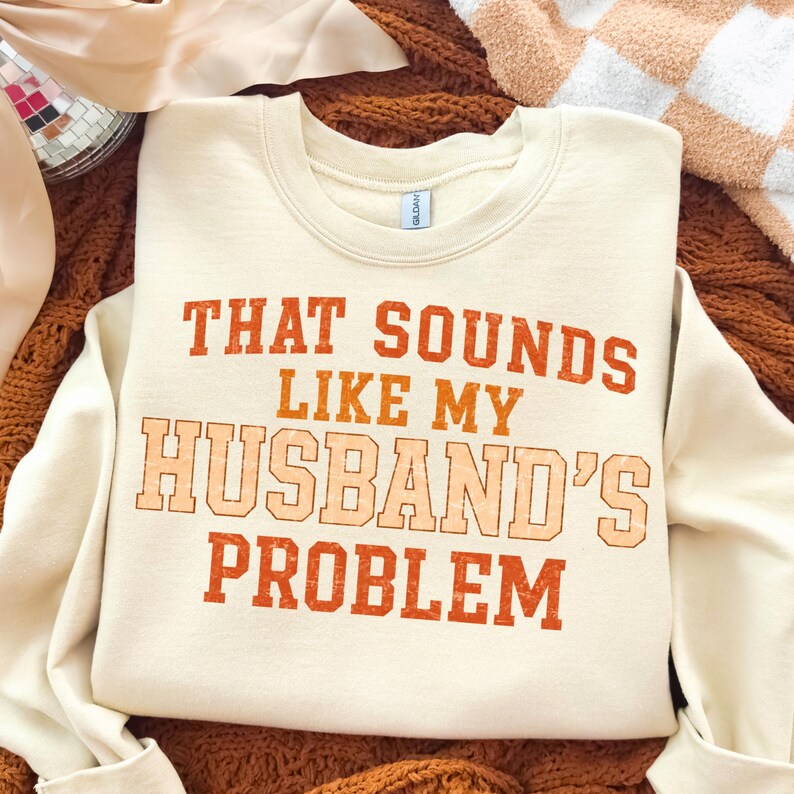 May include: Cream-colored sweatshirt with the text "THAT SOUNDS LIKE MY HUSBAND'S PROBLEM" in orange and peach lettering. The crew neck sweatshirt is made of a soft material and is displayed on a brown knitted surface.