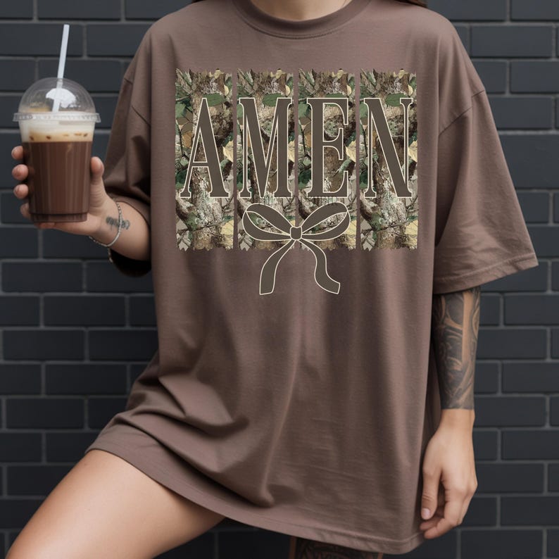 K&ouml;nnte beinhalten: Ein braunes T-Shirt mit dem Wort "AMEN" in einem Camouflage-Muster und einer Schleifen-Grafik. Das Shirt hat eine einfarbige braune Farbe und kurze &Auml;rmel. Die Person h&auml;lt einen transparenten Plastikbecher mit einem braunen Getr&auml;nk und einem Strohhalm.