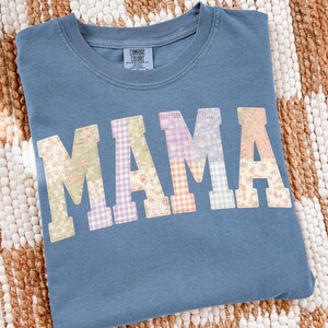 May include: A blue Comfort Colors sweatshirt with the word "MAMA" in patchwork letters. Each letter features a different floral or gingham pattern in shades of green, purple, and peach. The sweatshirt is displayed on a woven, cream-colored surface.