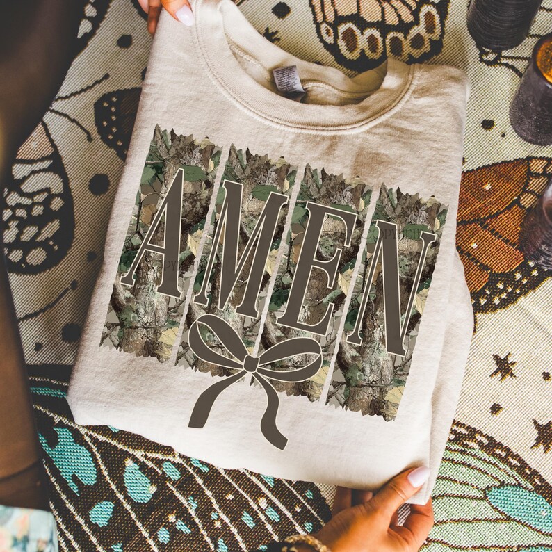K&ouml;nnte beinhalten: Ein beiges Sweatshirt mit dem Wort "AMEN" in brauner Schrift, vor einem Camouflage-Hintergrund. Unter dem Wort befindet sich eine braune Schleife. Das Sweatshirt wird von einer Person gehalten.