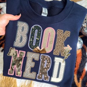 May include: Navy blue sweatshirt with the words "BOOK NERD" in a patchwork design. The letters feature floral, leopard print, and book page patterns. A butterfly and a small open book are also included in the design.
