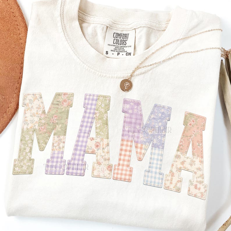 May include: Cream-colored t-shirt with the word "MAMA" in patchwork letters. The letters feature floral and gingham patterns in pastel colors. A gold necklace with a pendant rests on the shirt.