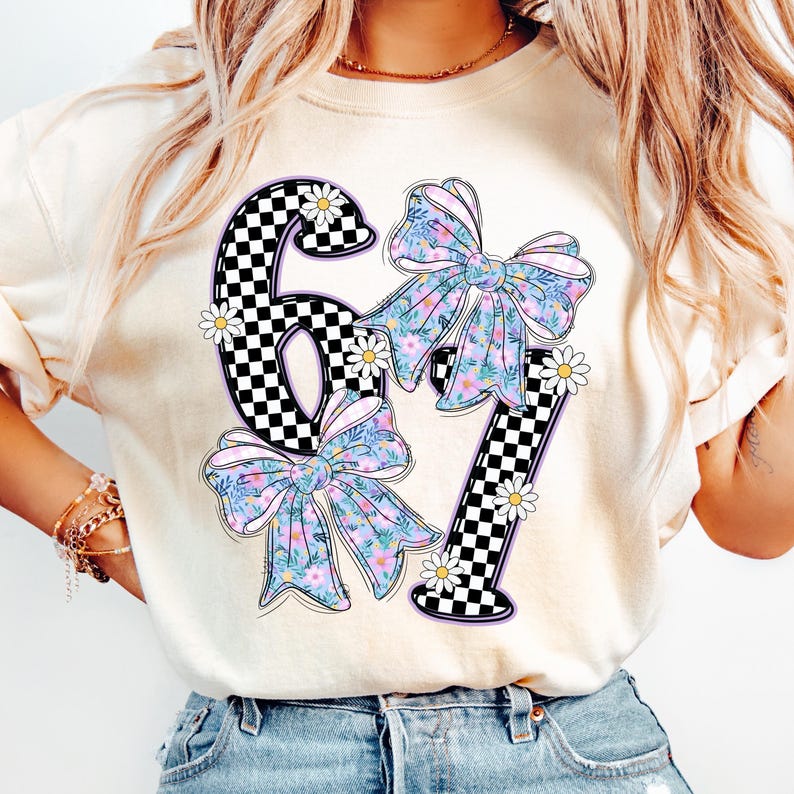 May include: Cream-colored t-shirt featuring the number "67" in a black and white checkered pattern. The numbers are adorned with floral bows in shades of blue, purple, and green, and accented with small white daisies.