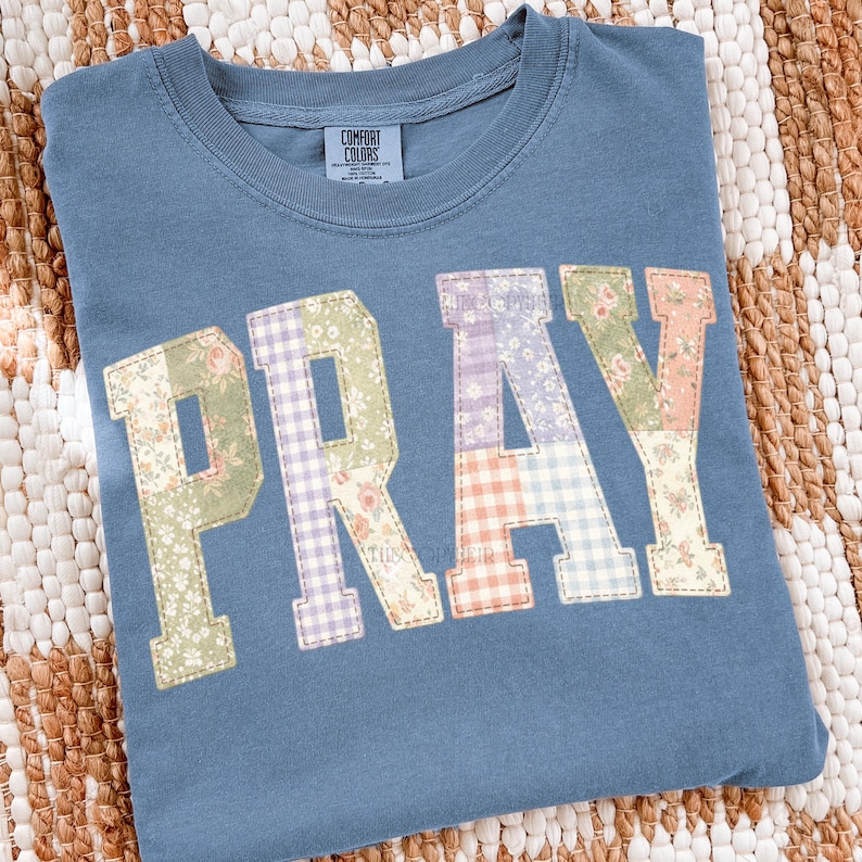 K&ouml;nnte beinhalten: Ein blaues Comfort Colors Sweatshirt mit dem Wort "PRAY" auf der Vorderseite. Die Buchstaben bestehen aus Stoff mit Blumen- und Vichy-Muster in Gr&uuml;n-, Lila- und Pfirsicht&ouml;nen.