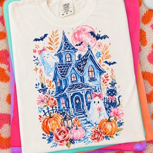 May include: An off-white t-shirt featuring a colorful graphic print of a blue haunted house, two ghosts with pink bows, a black cat, orange pumpkins, pink flowers, and bats under a large pink moon. The shirt has a Comfort Colors brand neck label.