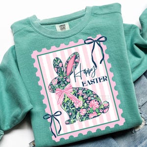 May include: A teal Comfort Colors sweatshirt featuring a pink and white striped postage stamp design with a floral bunny and the words "Happy Easter". The bunny has a pink bow and ears. Navy blue bows are on the top and bottom of the stamp.