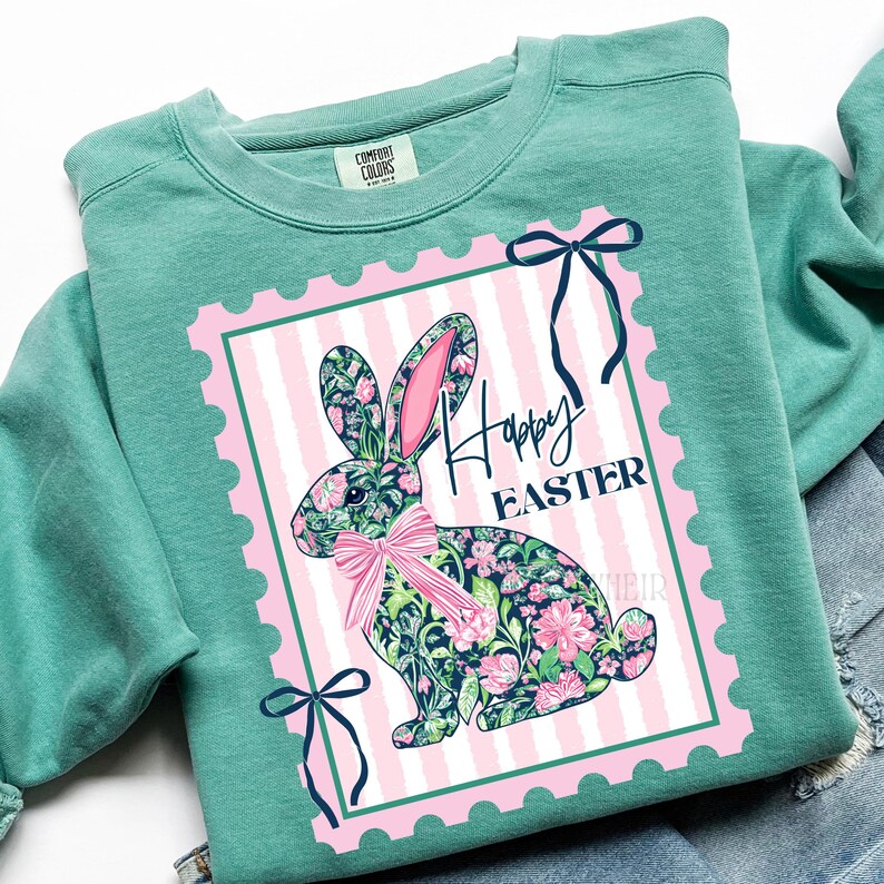 May include: A teal Comfort Colors sweatshirt featuring a pink and white striped postage stamp design with a floral bunny and the words "Happy Easter". The bunny has a pink bow and ears. Navy blue bows are on the top and bottom of the stamp.