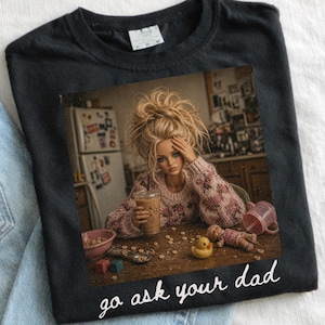 Pu&ograve; includere: T-shirt nero con un'immagine di una bambola che tiene una bevanda al caff&egrave;, circondata da giocattoli e cereali. Il testo "go ask your dad" &egrave; stampato in bianco sul fondo della maglietta.